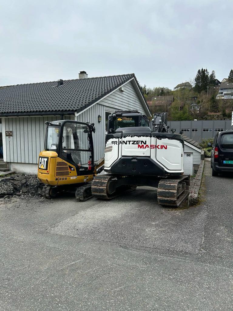 To gravemaskiner parkert foran en bolig, asfalt og steinarbeid pågår.