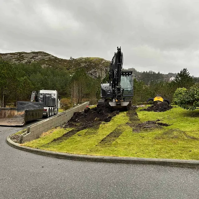 Gravemaskin jobber på grønn plen, lastebil står parkert ved murkant.