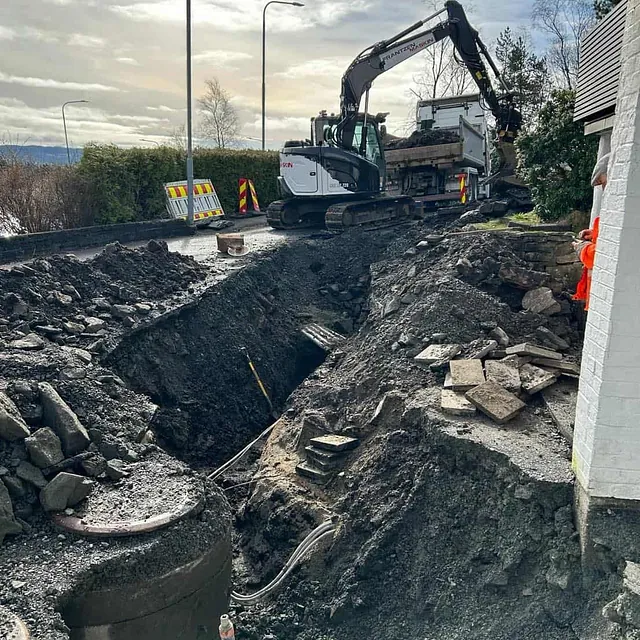 Større utgraving ved bolig og vei, gravemaskin laster masse i lastebil.