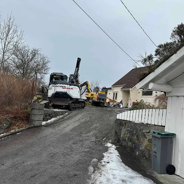 Gravemaskiner parkert i oppoverbakke mellom bolighus og steinmur.
