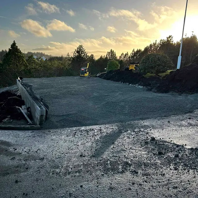 Planert tomt med grus, maskiner og trær i bakgrunnen, solnedgang.